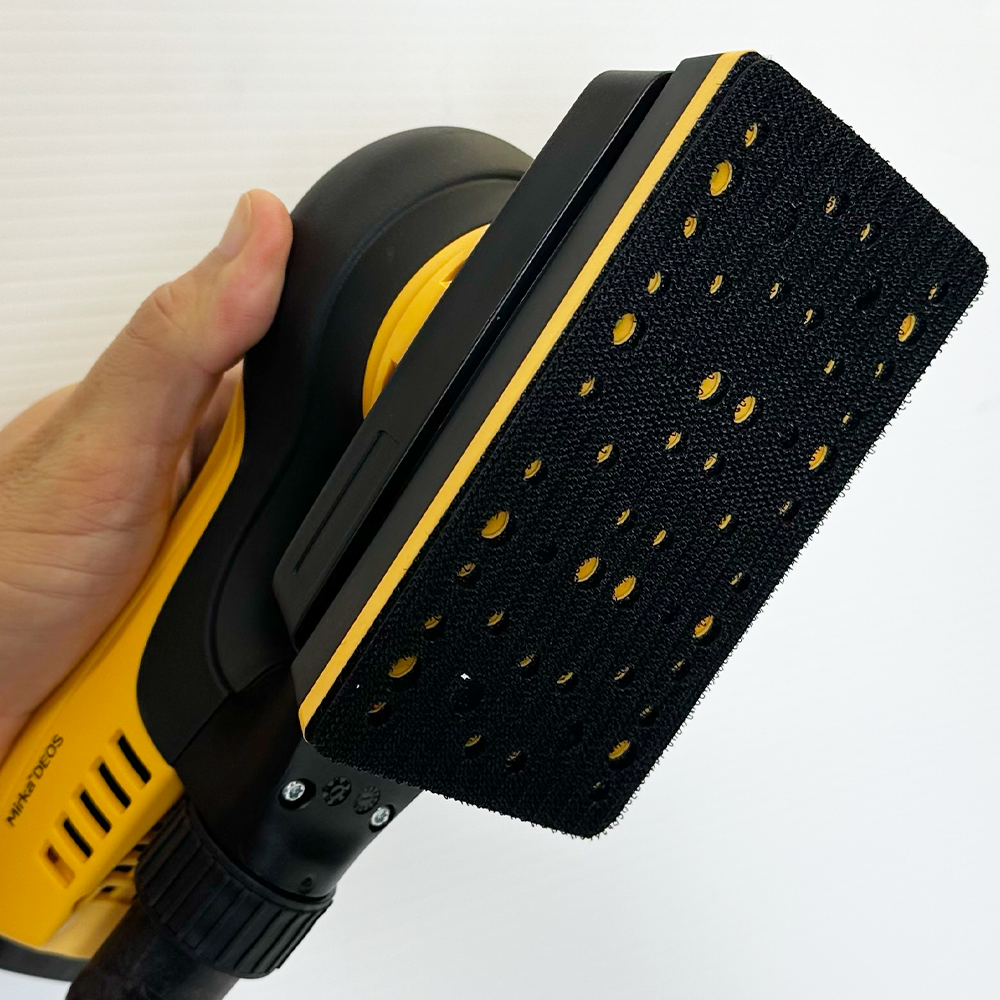 Hand holding a Mirka DEOS sander with a black 81x133mm pad saver fitted to the base